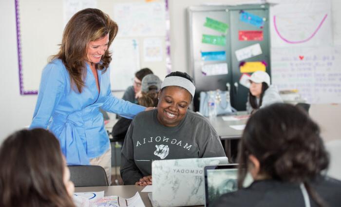 A teacher in a blue blouse smiles and places a hand on a student's shoulder. The student, wearing a gray "Magnolia" sweatshirt, smiles at her laptop. Other students work in the background.